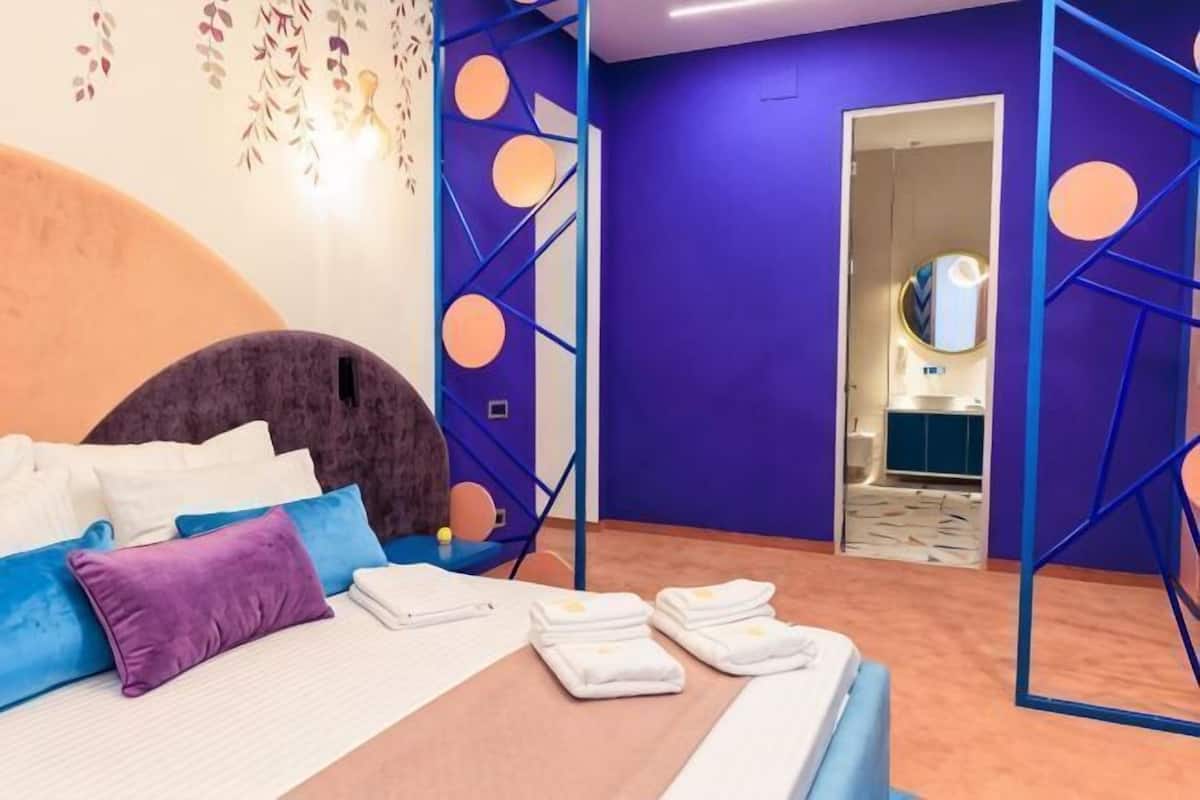 A bedroom with a bed, pillows, a bedside table, a mirror, and a bathroom door.