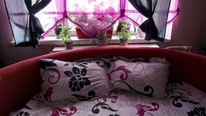 Una cama con ropa de cama de estampado floral, una ventana con plantas en macetas y cortinas translúcidas recogidas.