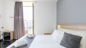 A hotel room with a large bed, a wooden headboard, a view of the city through the window, and a small bedside table with a lamp. - Palace Napoli (Naples)