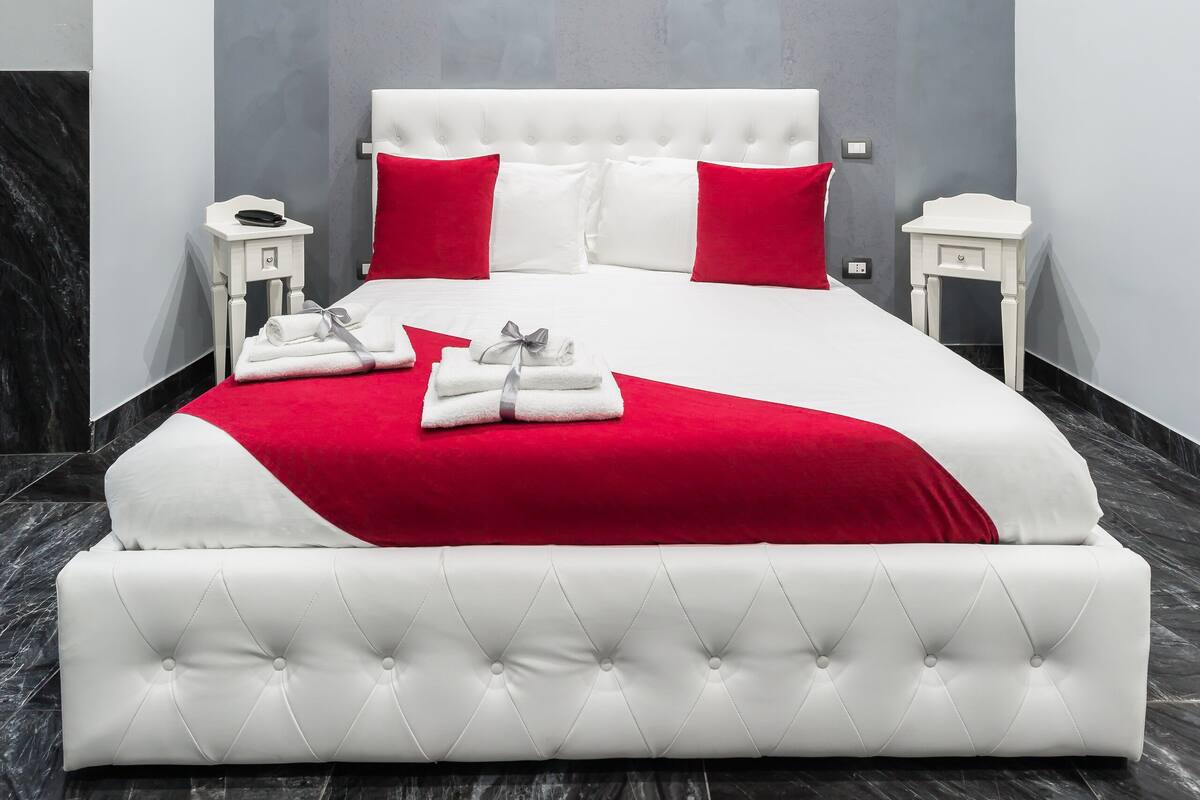 A modern bedroom with a white tufted headboard, red and white bedding, and marble flooring.