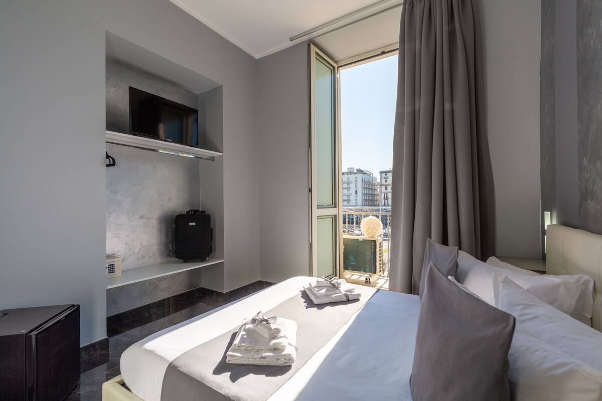 A modern hotel room with a bed, a small seating area, a TV, and a view of the city.