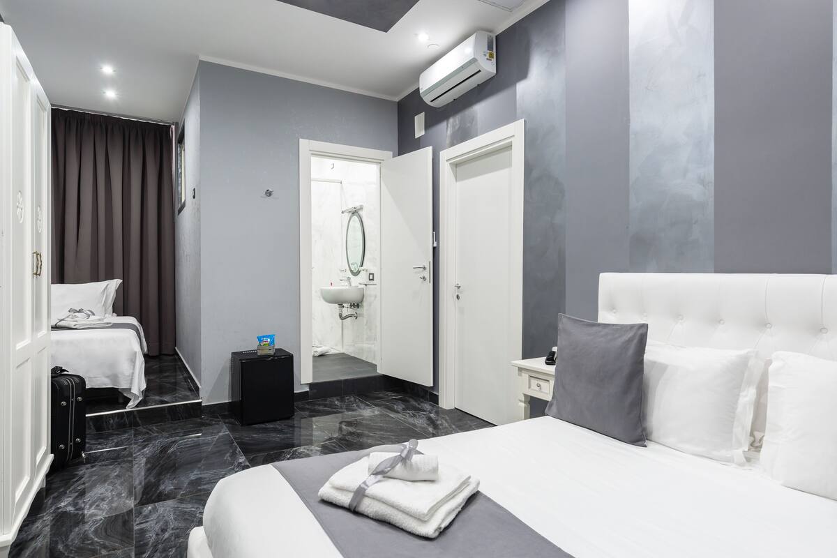 A modern hotel room with a large bed, a smaller bed, a bathroom with a sink, and a suitcase on the floor.