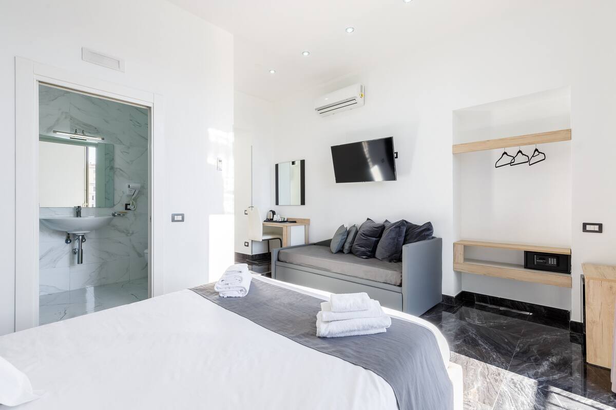 A modern hotel room with a large bed, a built-in sink, a flat-screen TV, and a seating area with a sofa and pillows.