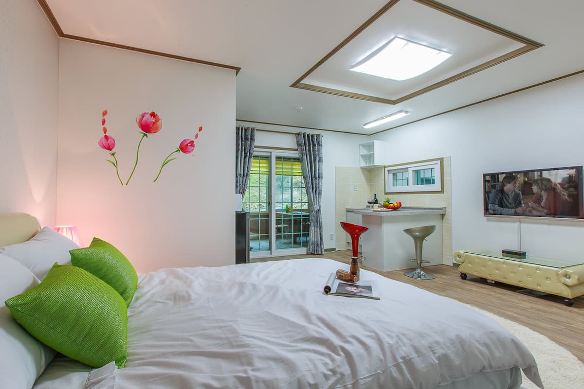 A bedroom with a bed, green pillows, a window with curtains, a bathroom with a sink, and a TV.