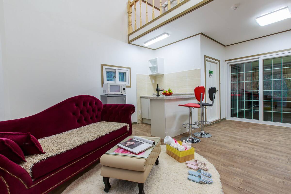 A living room with a red sofa, a glass coffee table, a small ottoman, a kitchen area with a microwave, a red bar stool, and a large window with a view.