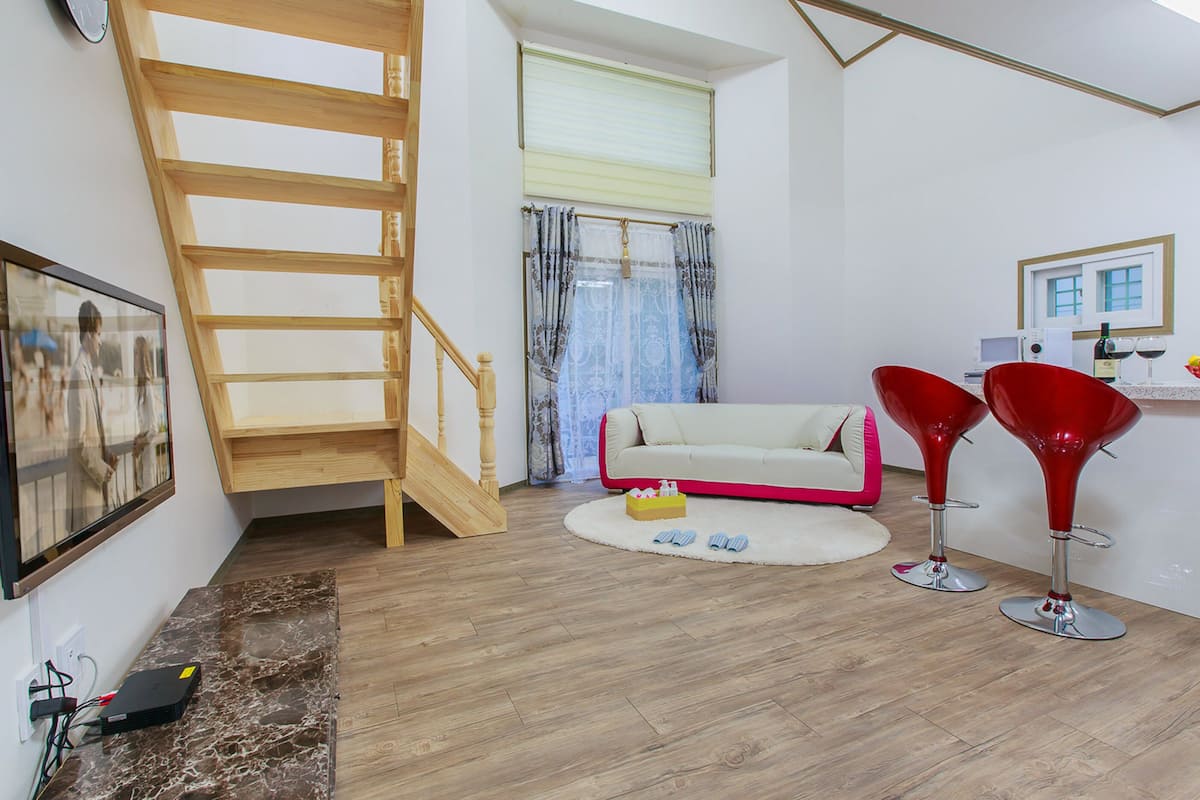 A modern living room with a wooden staircase, a white sofa, two red chairs, a small rug, and a television mounted on the wall.