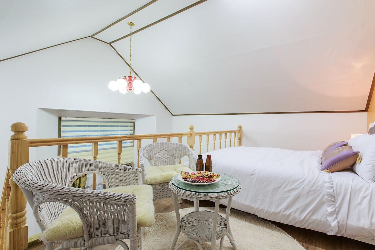 A cozy bedroom with a bed, wicker chairs, a small table with a plate of food, and a hanging light fixture.
