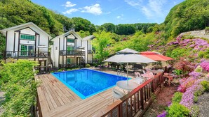 A pool with a wooden deck, surrounded by lush greenery and a hillside. - Sky Wind and Star SPA Pension (Pocheon)