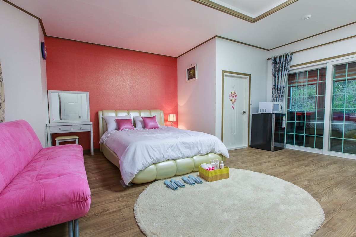 A bedroom with a bed, a pink chair, a white bedside table, a mirror, a white door, and a window with curtains.