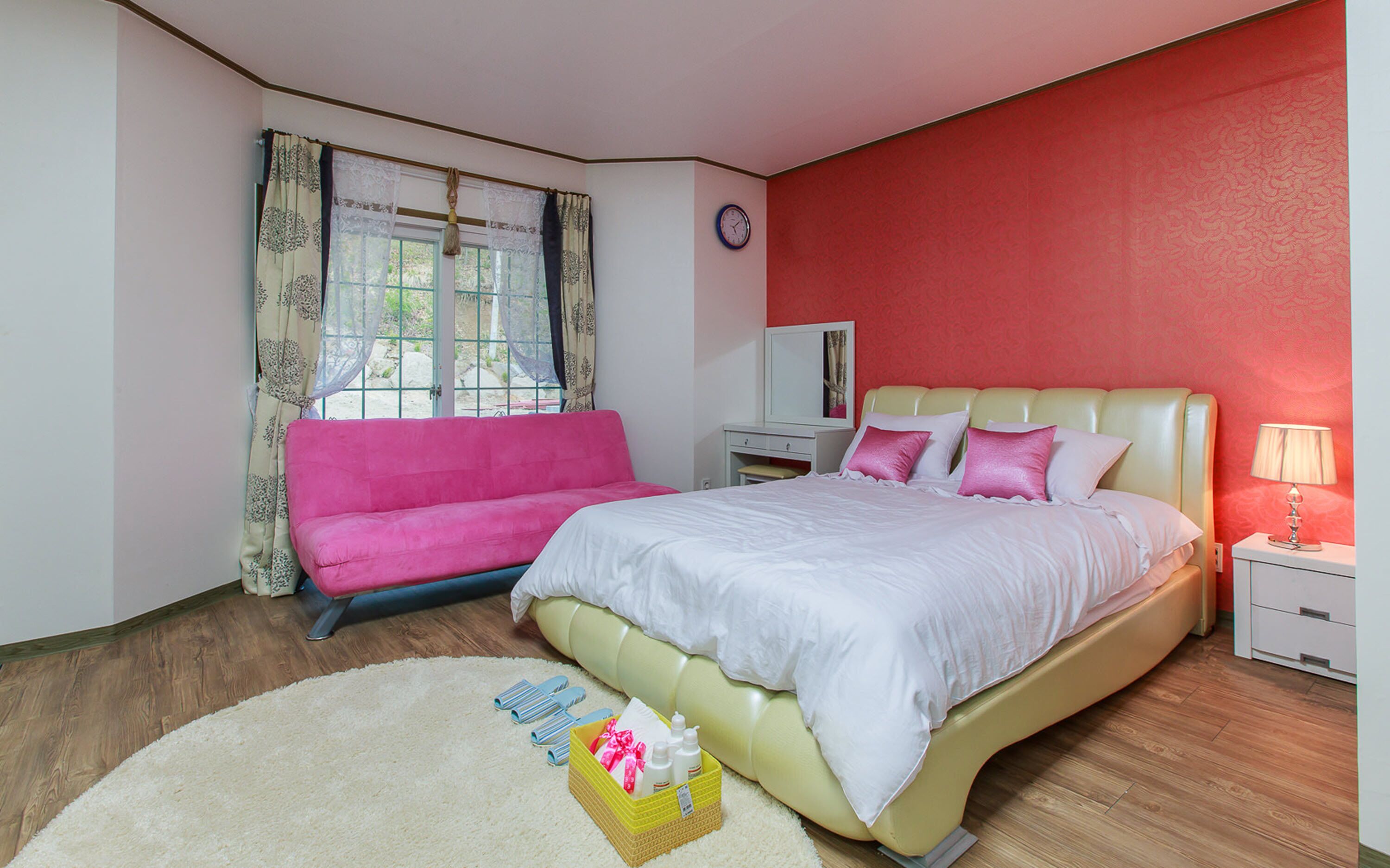 Un dormitorio con cama, un sofá rosa, una mesita de noche, una lámpara y una ventana con cortinas.