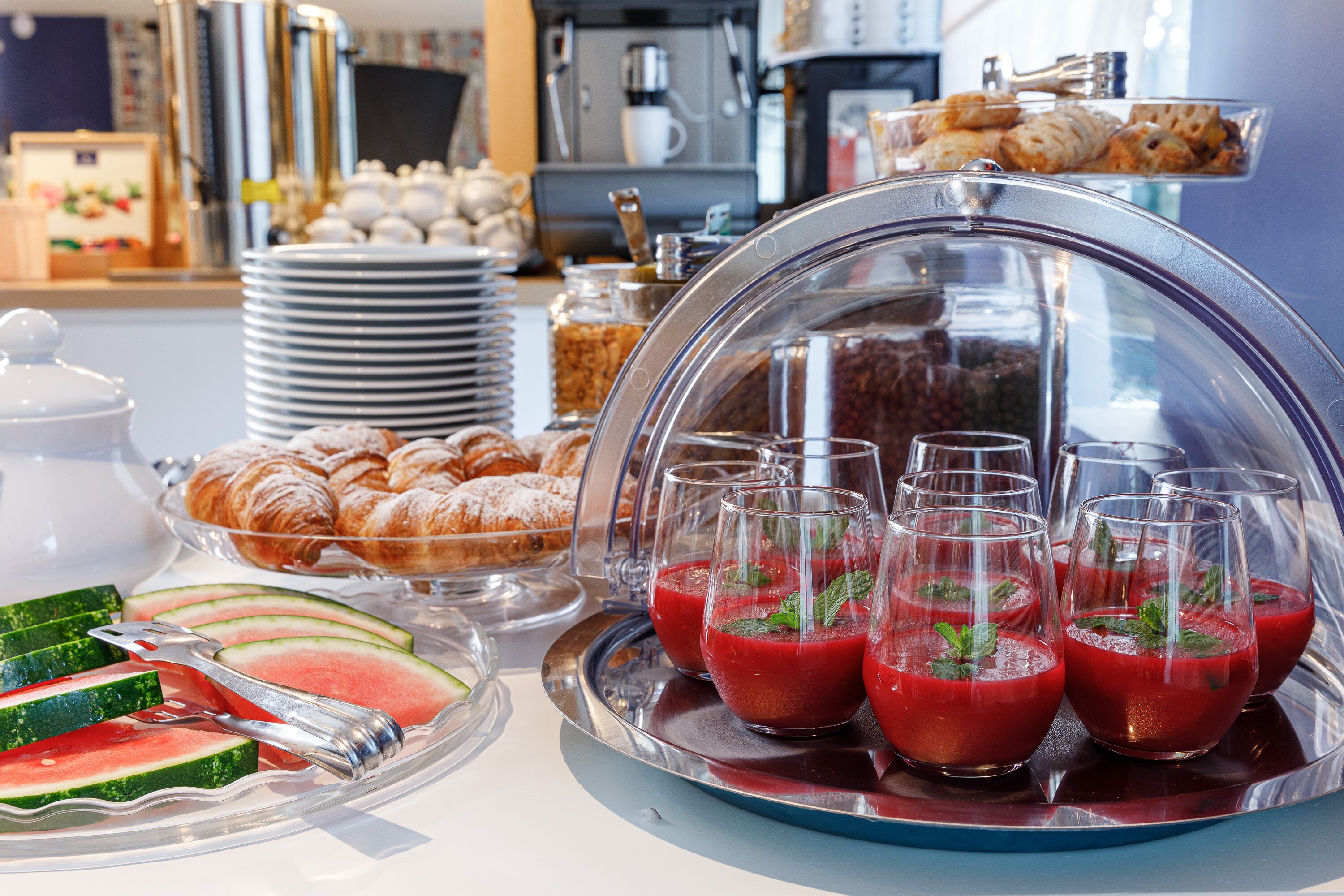 A breakfast buffet with sliced watermelon, croissants, and red juice glasses.