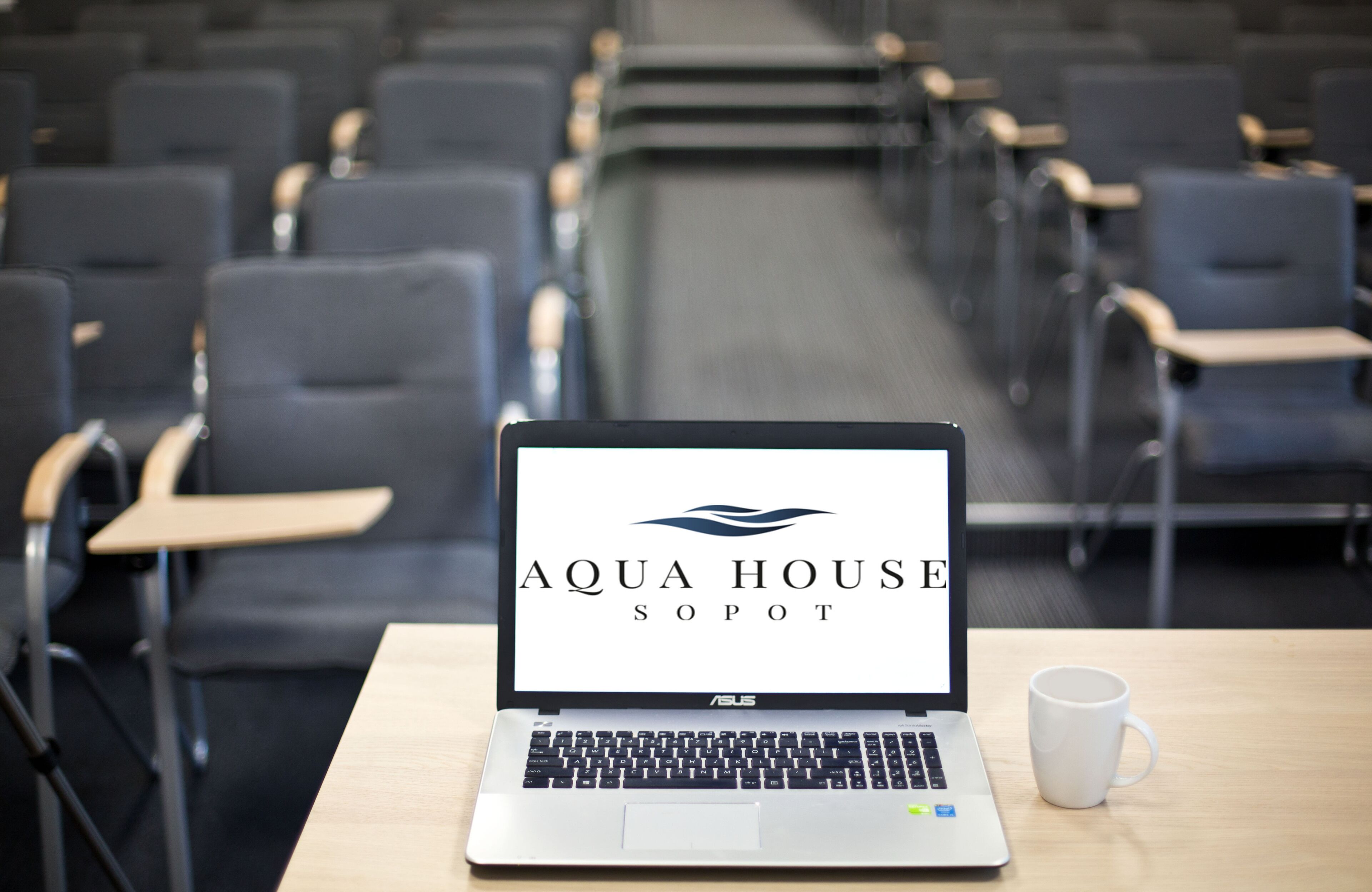 Una computadora portátil sobre una mesa con una taza blanca, mostrando el logotipo de Aqua House Sopot, en una sala de conferencias con filas de sillas.