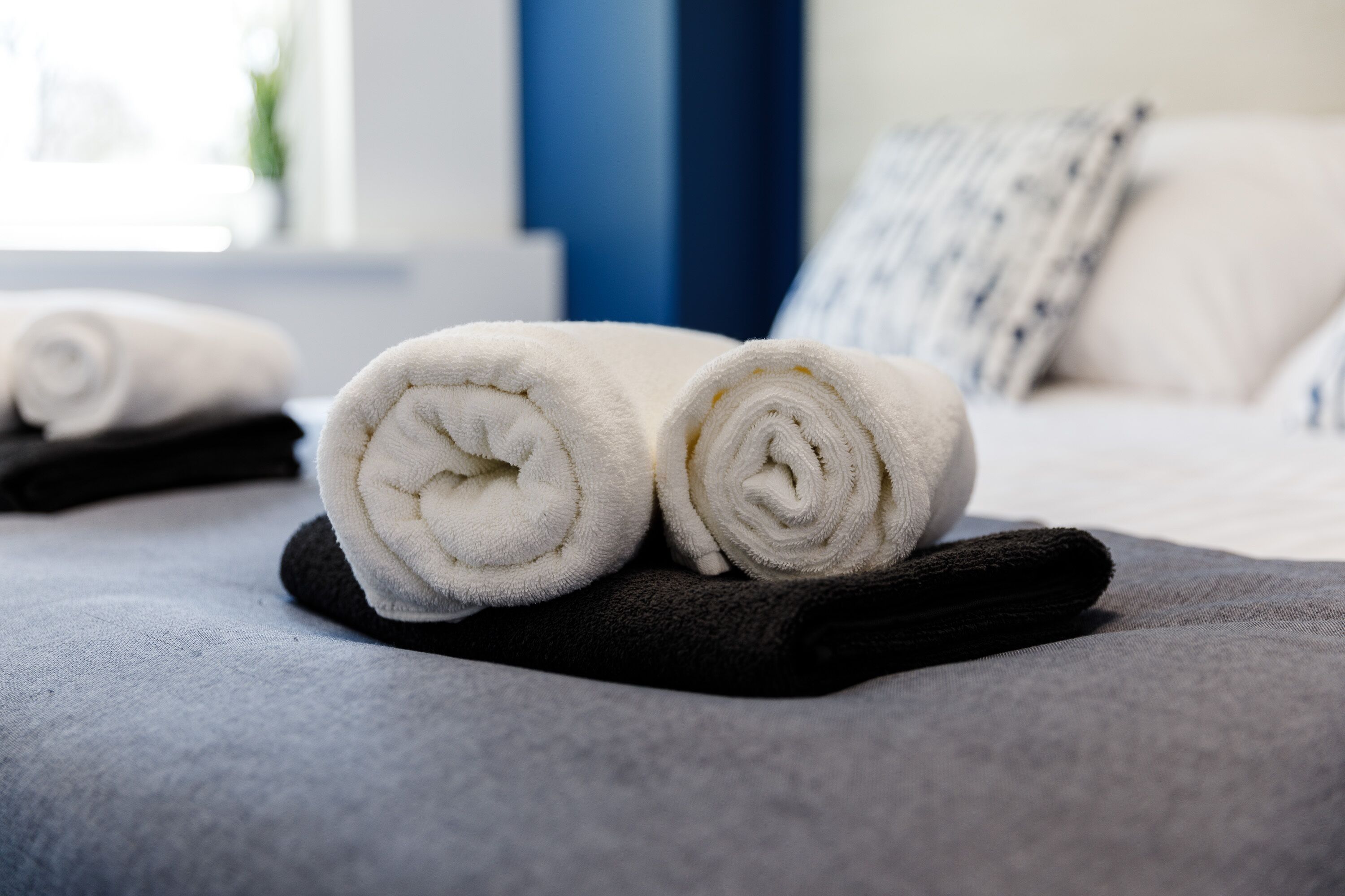 A neatly made bed with rolled white towels and a black towel on a grey bedspread.