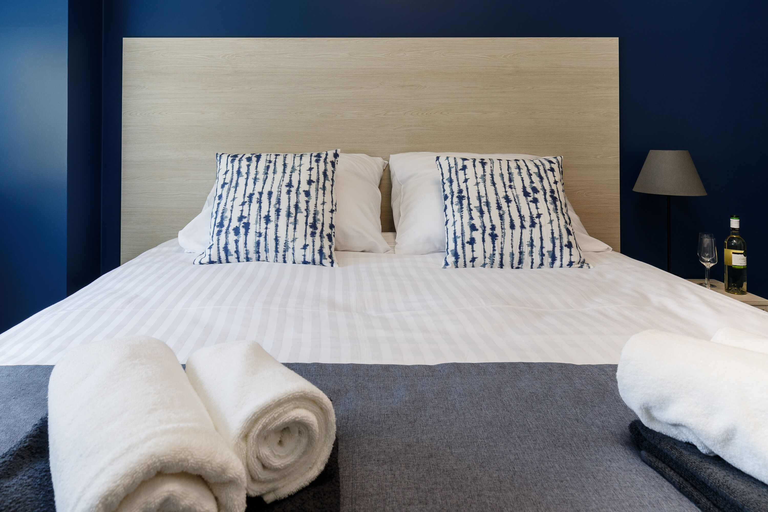 A neatly made bed with striped bedding and patterned pillows, a rolled white towel, and a grey blanket on a dark floor.