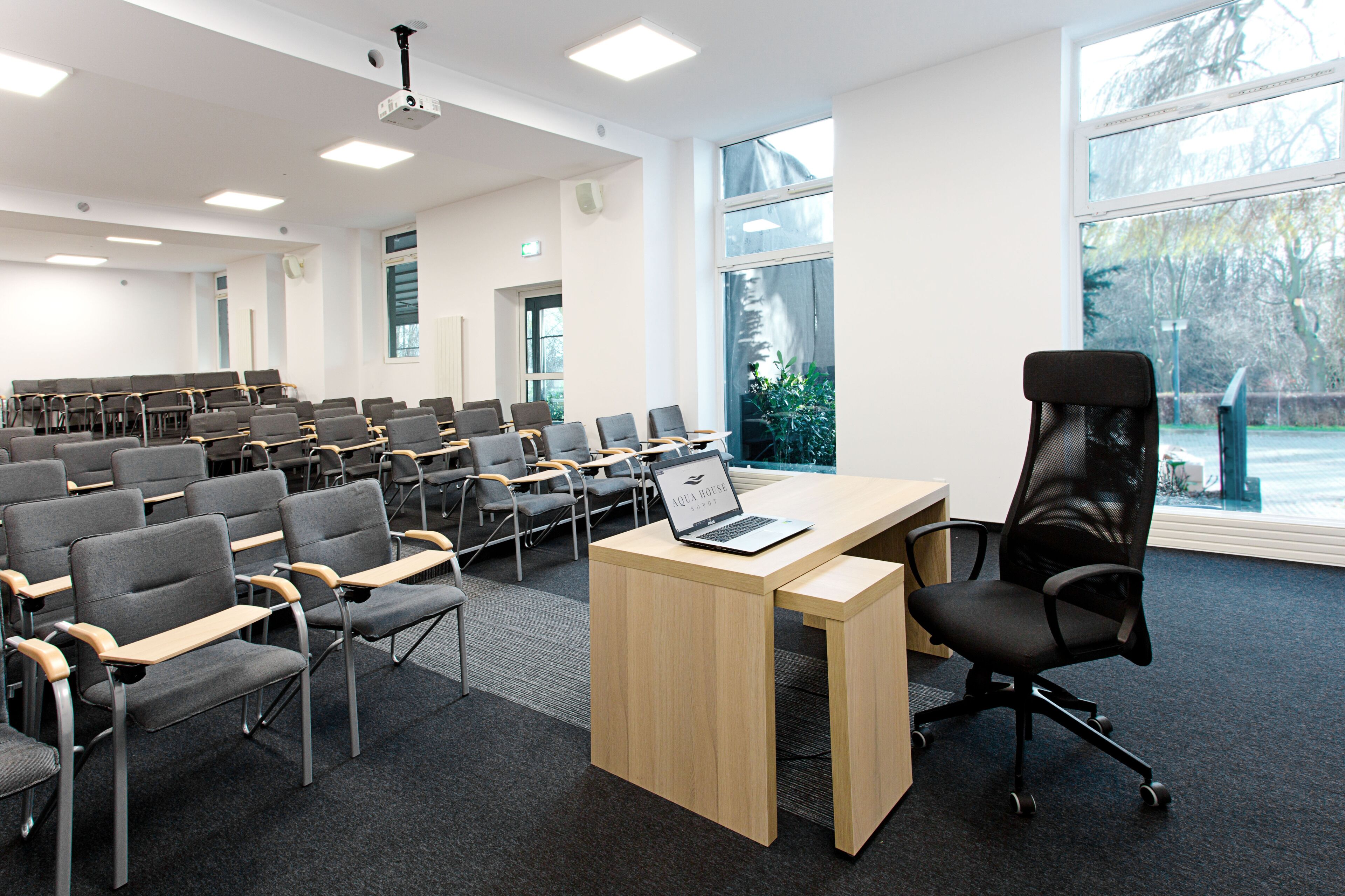 A conference room with rows of chairs, a wooden table with a laptop, and large windows with a view of trees.