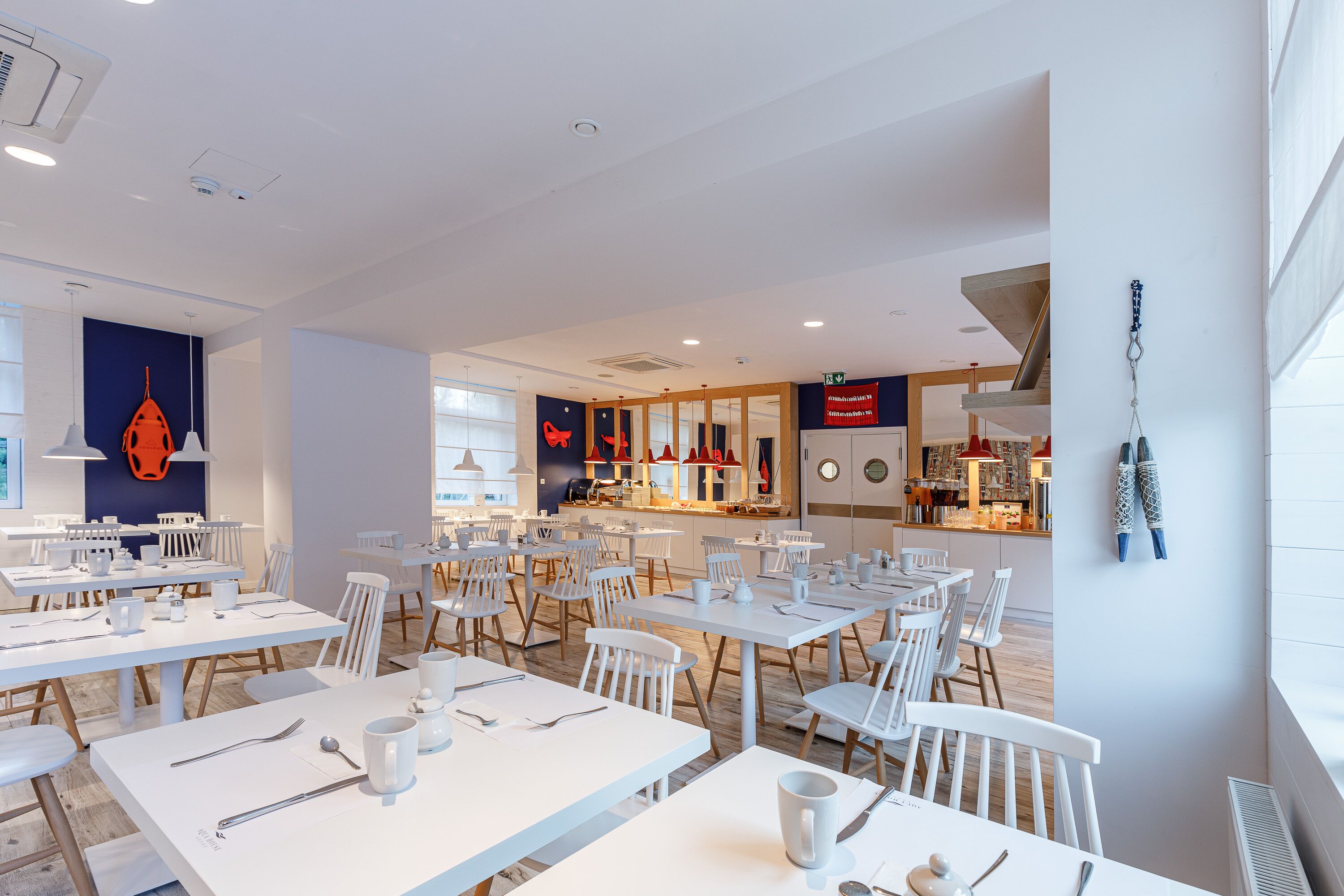 A modern dining area with white tables and chairs, a kitchen in the background, and a view of the city.