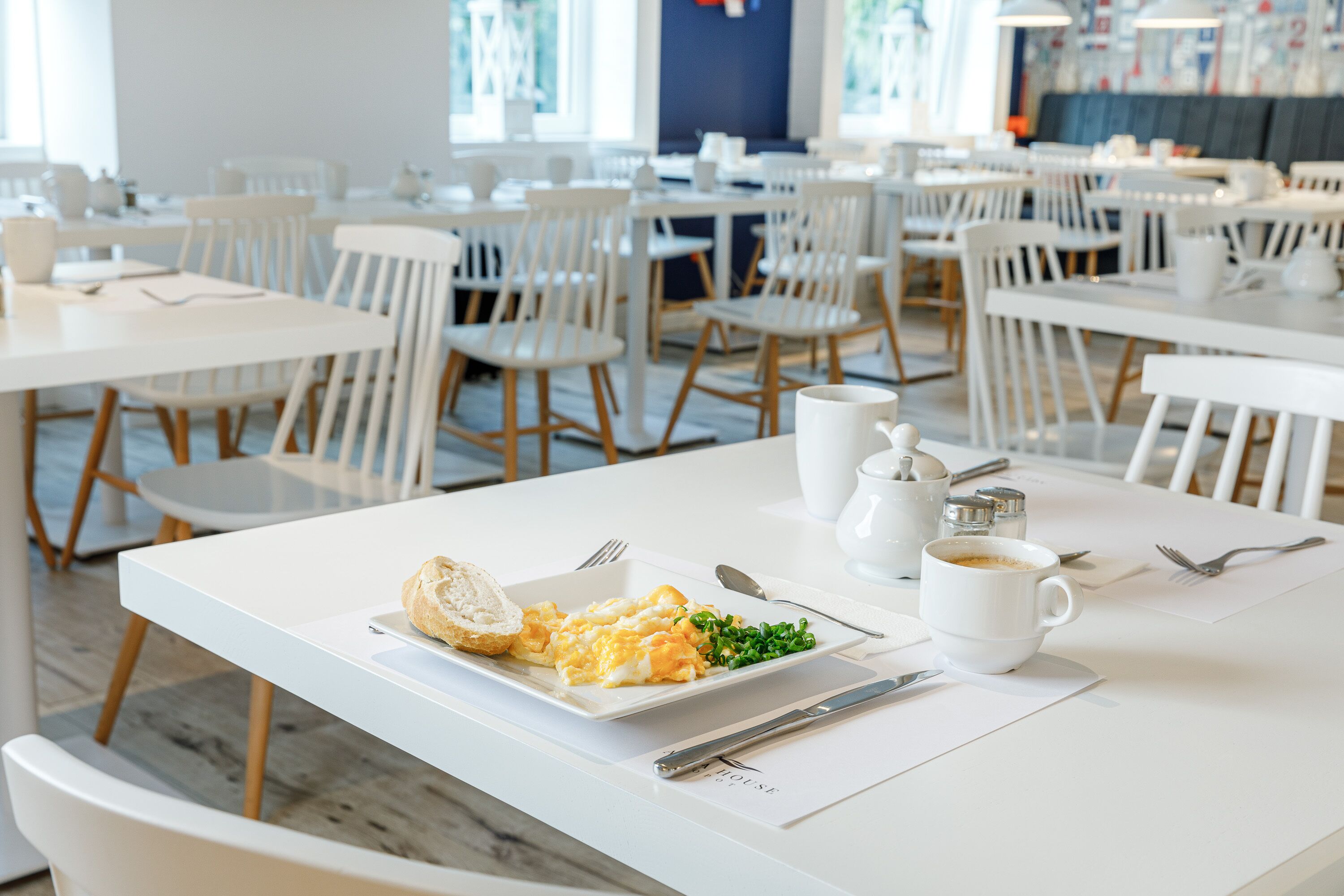 A clean, modern dining area with white tables and chairs, a plate of scrambled eggs, toast, and green vegetables.