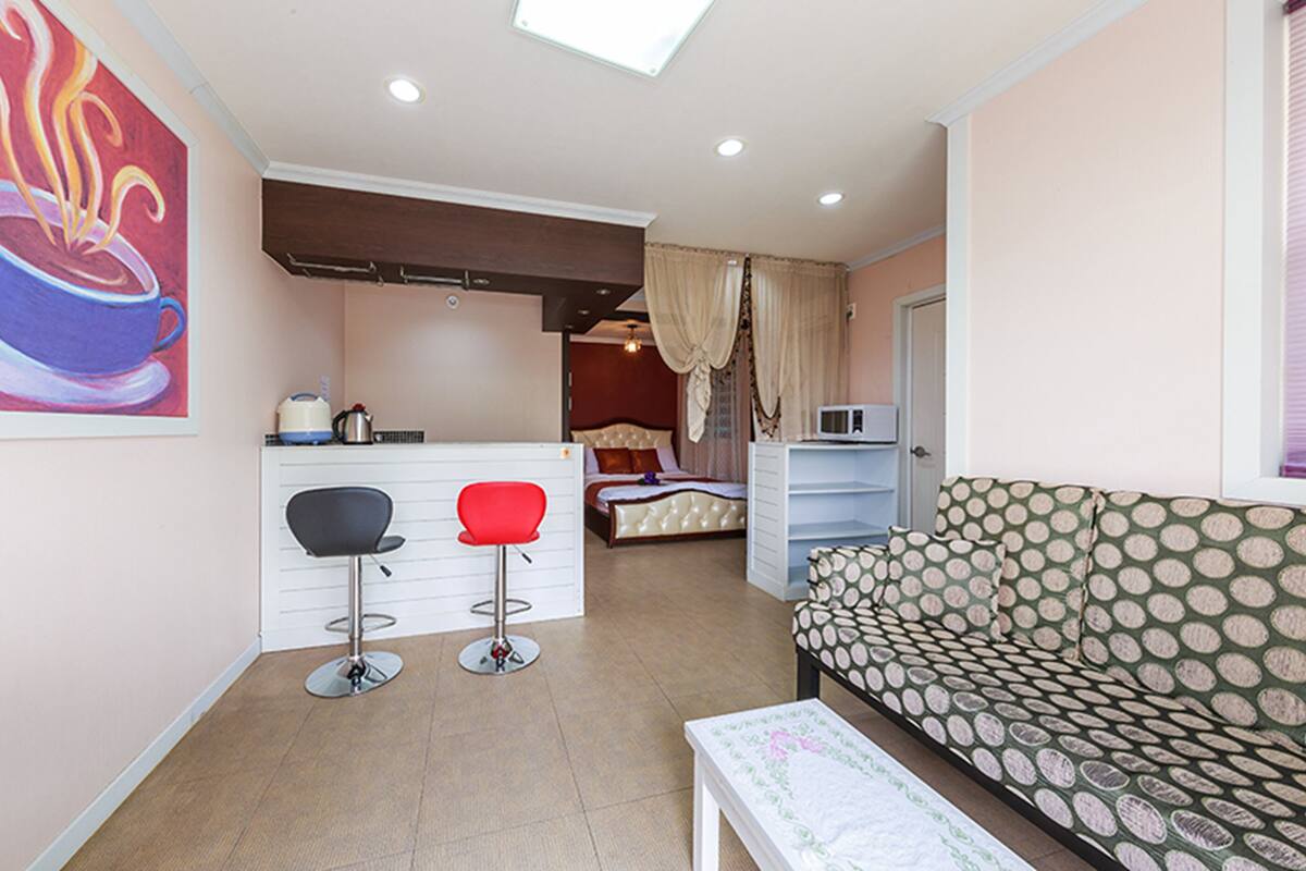 A compact living space with a kitchenette, a bed, a dining area with two stools, and a patterned sofa.