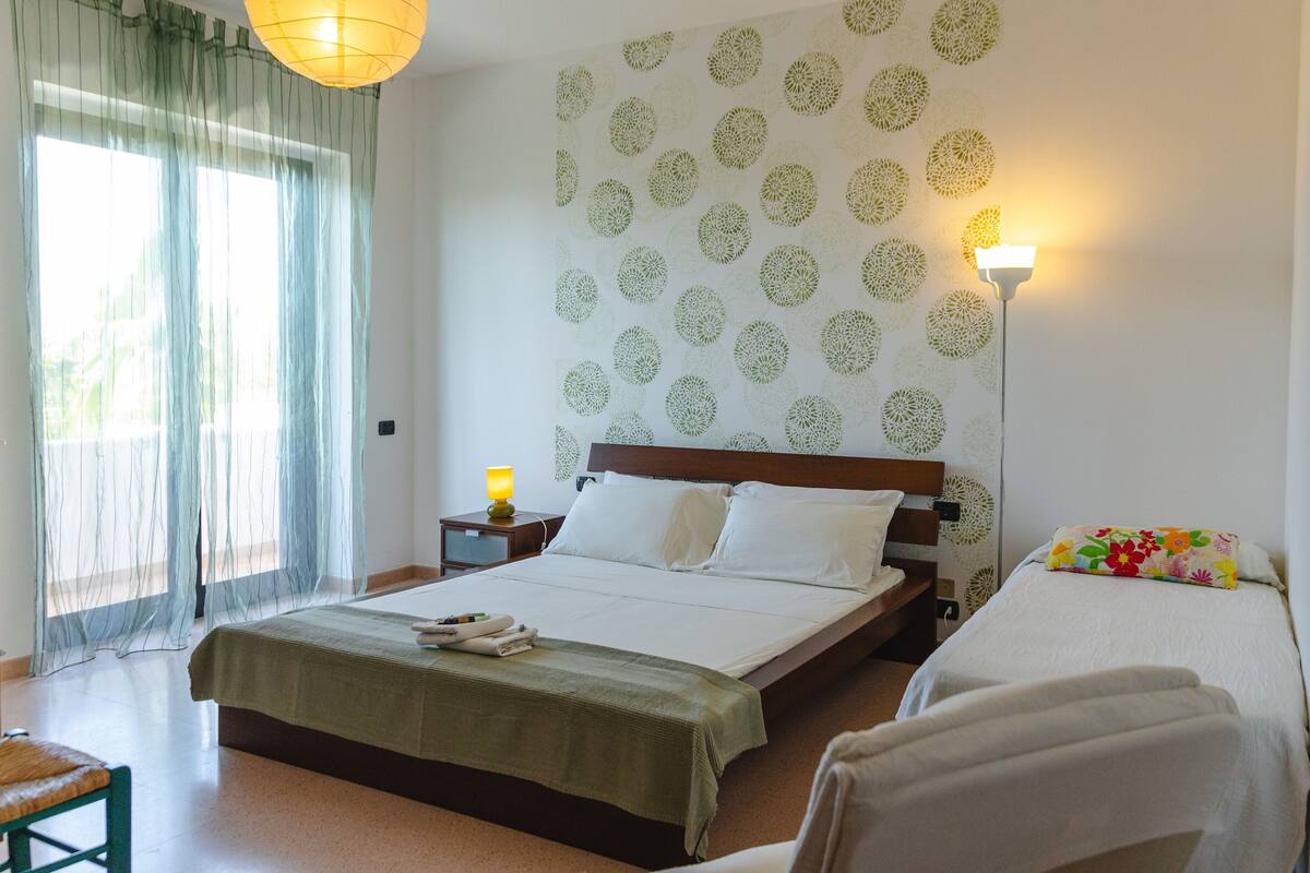 A hotel room with a bed, a bedside table with a lamp, a chair, and a window with sheer curtains.