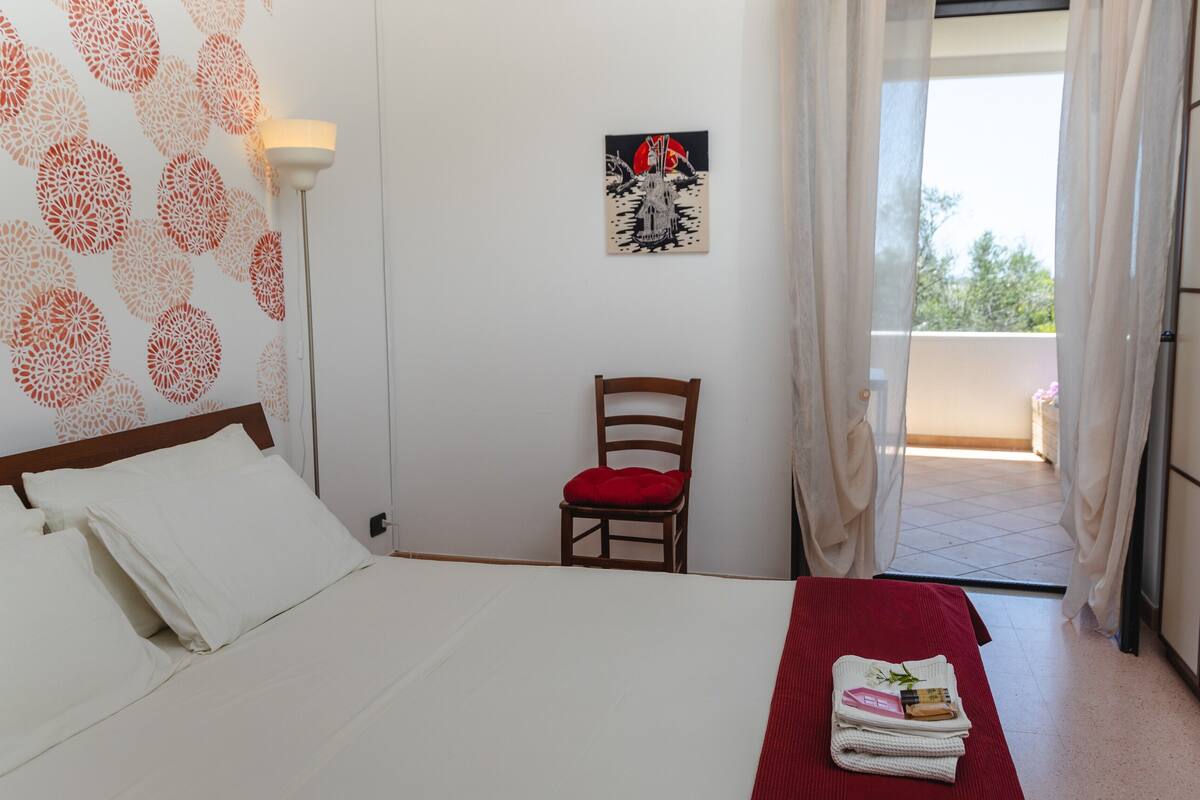 A hotel room with a bed, a chair, a small table with toiletries, and a balcony with a view.
