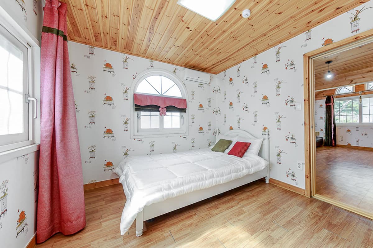 Ein Schlafzimmer mit Holzboden, einem Bett mit weißer Bettwäsche, einem Fenster mit rosa Vorhang und einem Spiegel, der den Raum reflektiert.