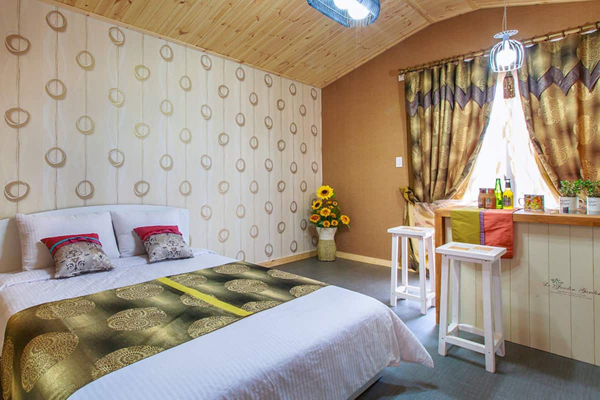 Ein Schlafzimmer mit einem Bett, Holzboden, einem Fenster mit Vorhängen, einem kleinen Tisch und einer Vase mit Blumen.