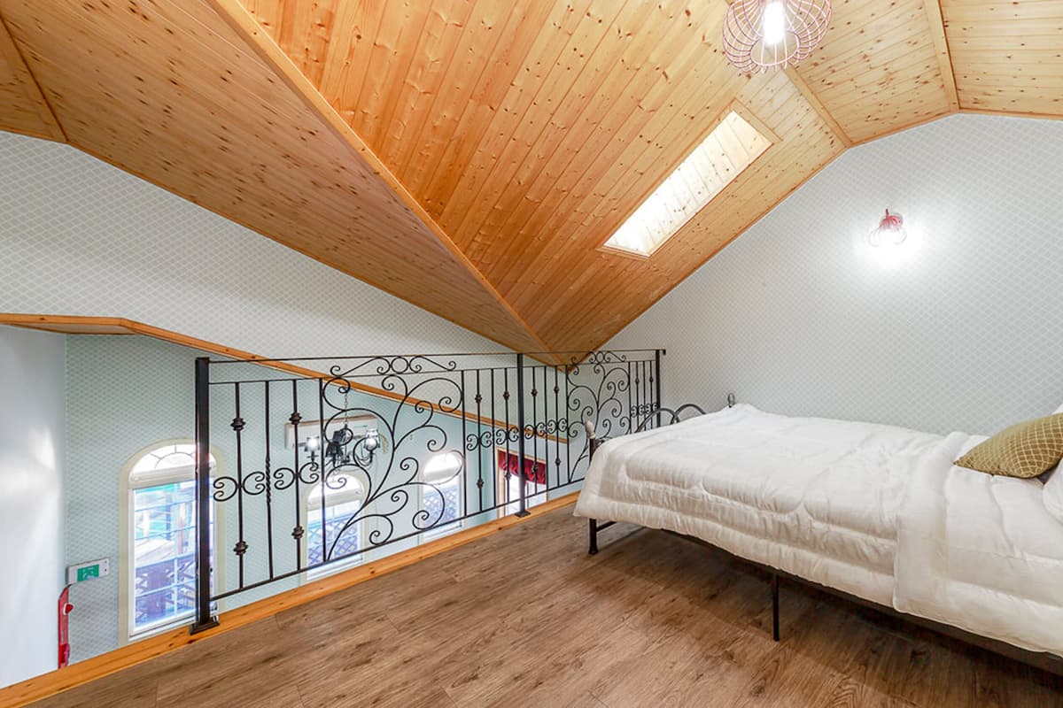 Ein Schlafzimmer mit schrägem Dach, einem Bett, einem Fenster und einem Balkongeländer.