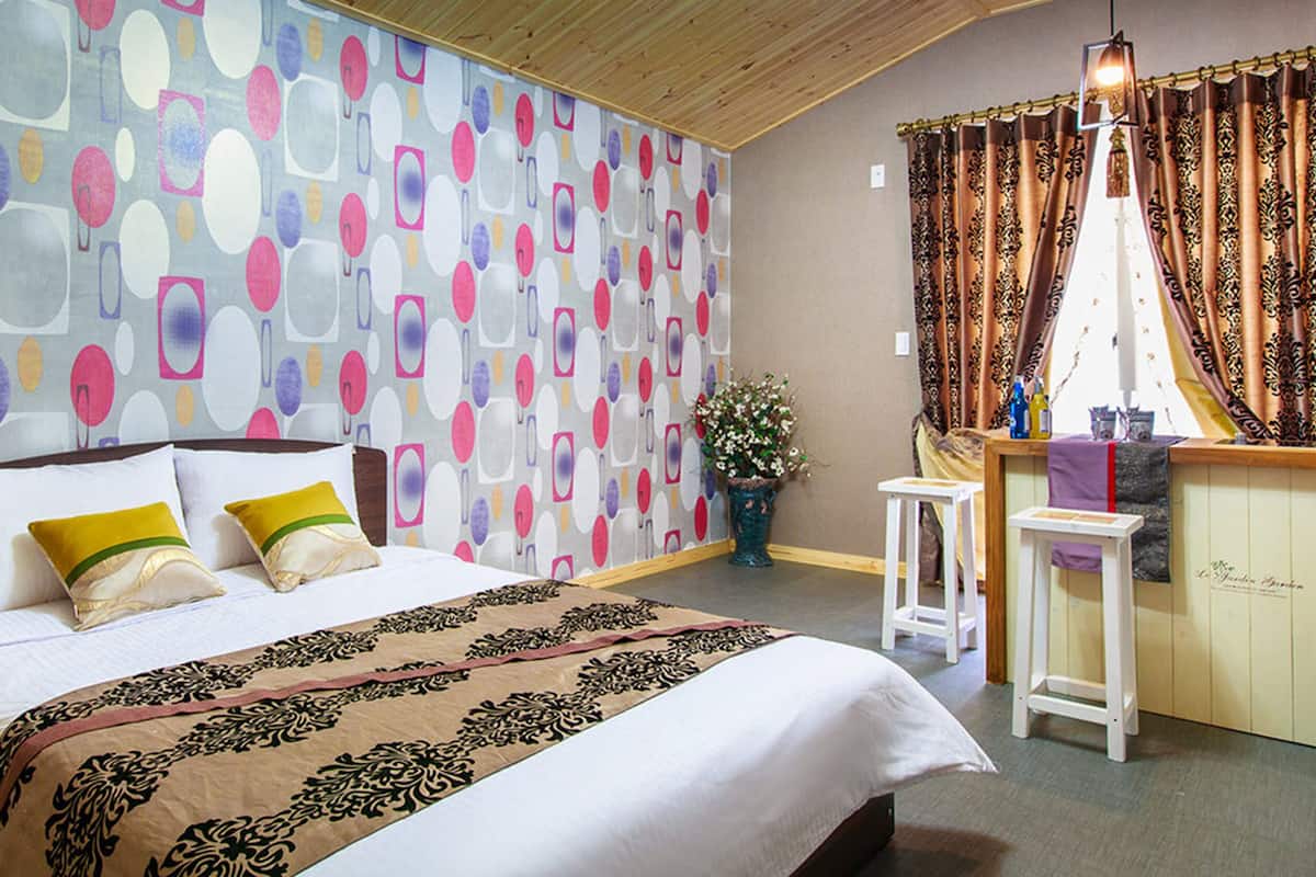 Ein Schlafzimmer mit Bett, Nachttisch, Stuhl, Fenster mit Vorhängen und Wand mit einem farbenfrohen abstrakten Muster.