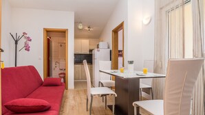 Un espacio habitable compacto con un sofá rojo, una mesa de comedor con sillas, una zona de cocina y una pequeña mesa con un vaso de jugo de naranja.