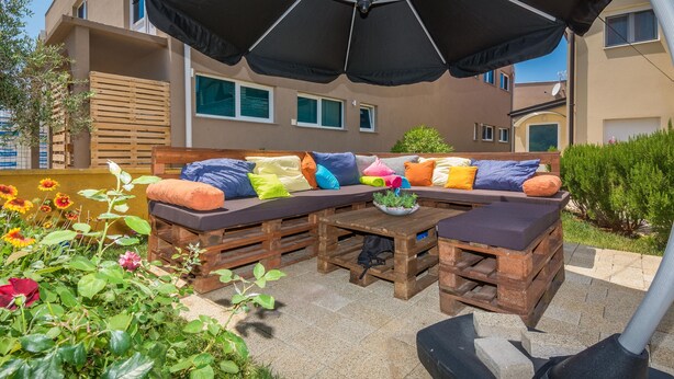 Un área de estar al aire libre con un banco y una mesa de madera, rodeada de plantas y flores en macetas.