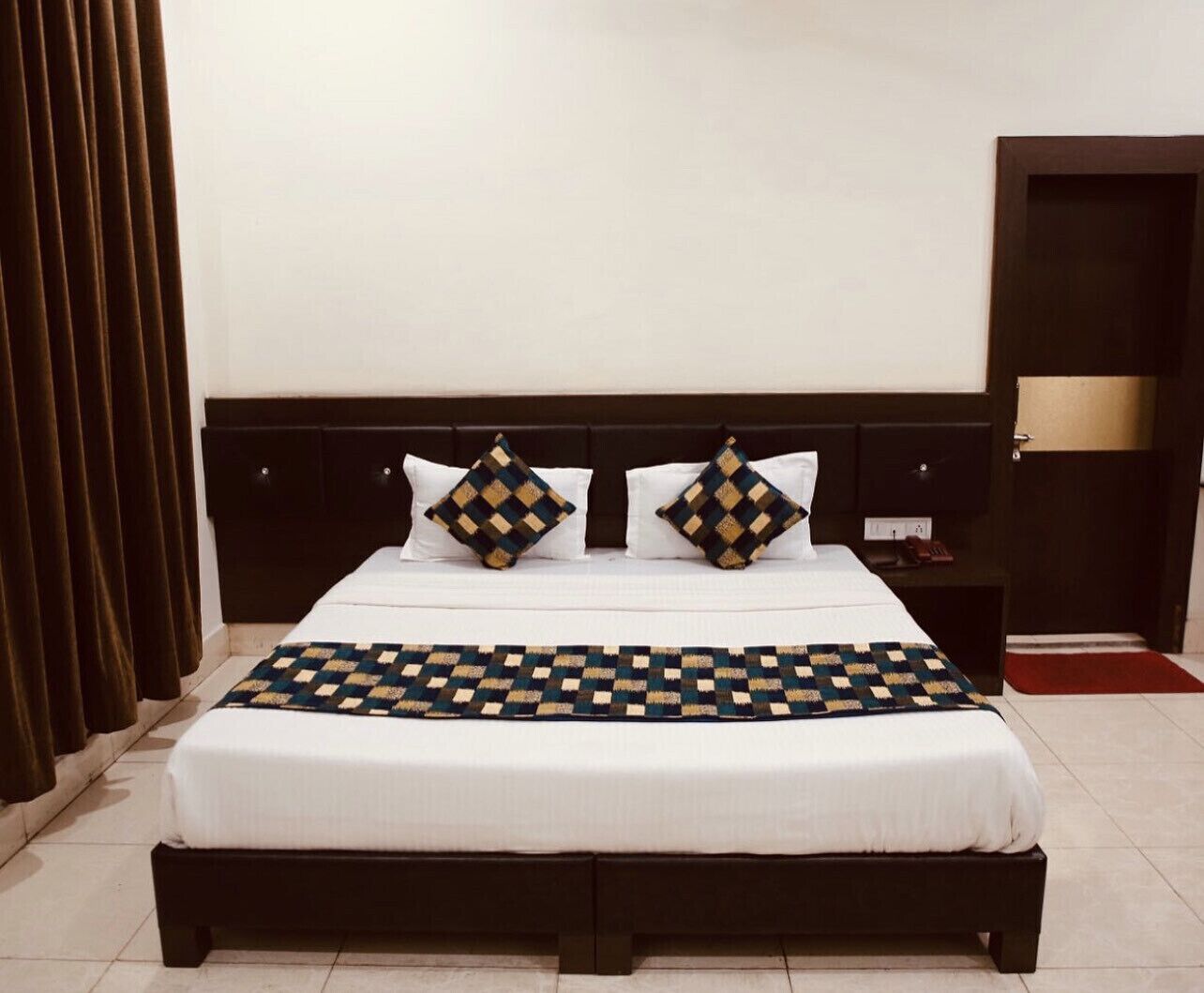 A neatly made bed with a checkered bedspread and two decorative pillows, a dark headboard, and a nightstand with a lamp.