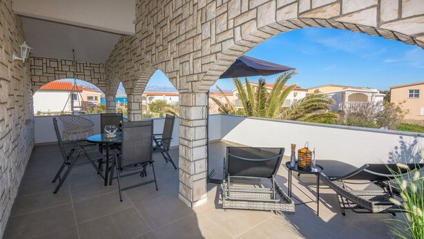 A covered outdoor area with stone pillars, a table with chairs, and a lounge chair. - Apartments Bojana (Vir)