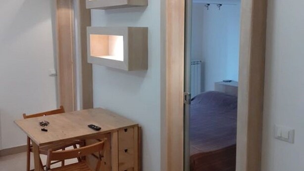 Il s’agit d’une pièce de taille modeste, comprenant un bureau en bois, une chaise et un lit. Des étagères intégrées sont fixées aux murs, et une porte donne accès à une autre pièce.