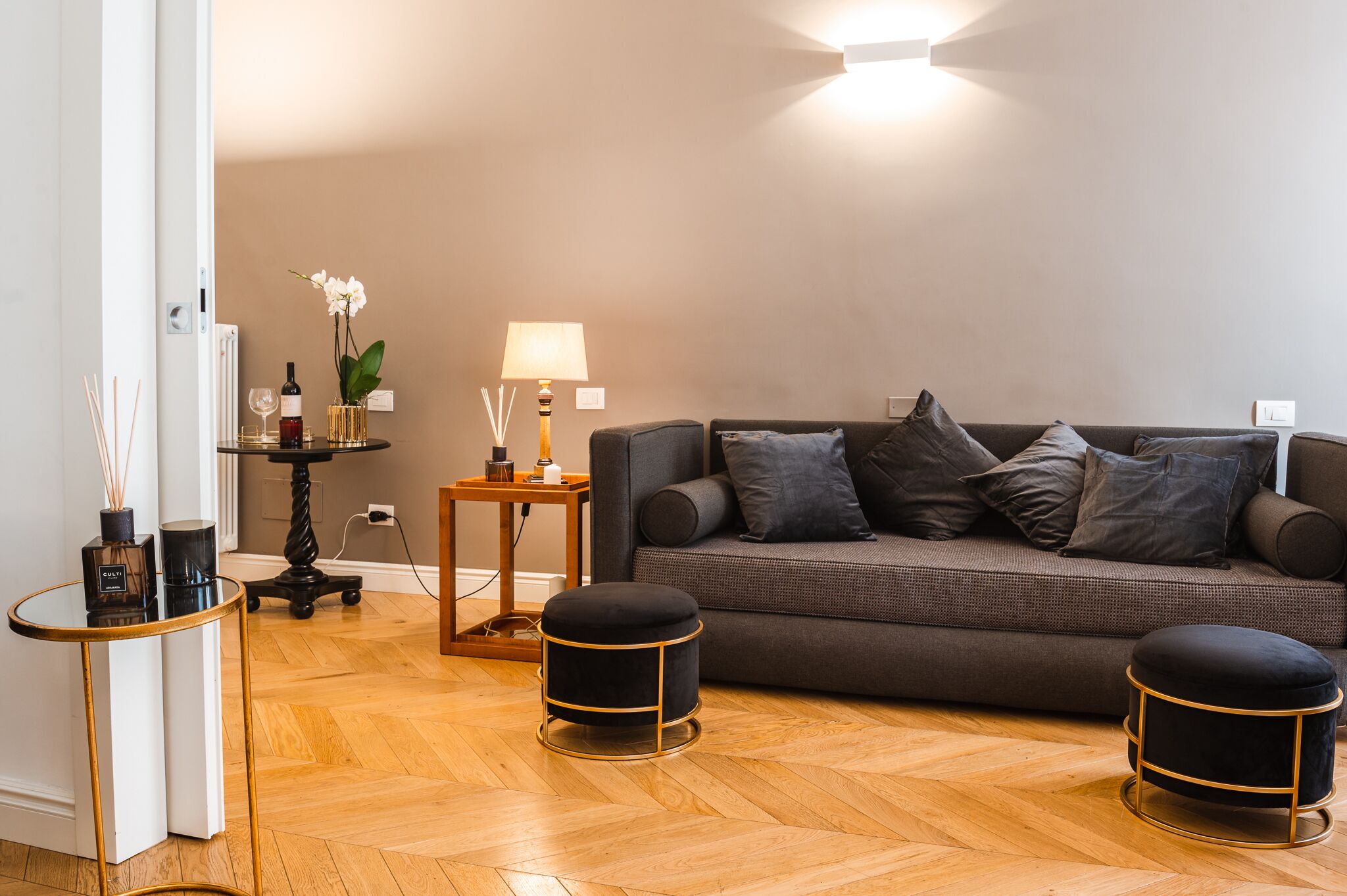 A modern living room with a grey sofa, two black ottomans, a small round table with a lamp, and a vase with an orchid.