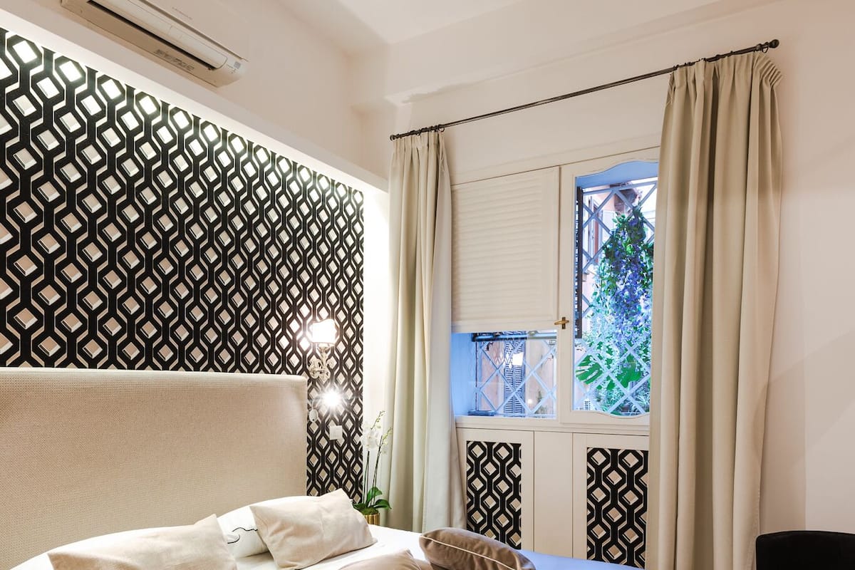 A neatly made bed with a beige bedspread and pillows, a patterned wall, a window with curtains, and a small table with a plant.