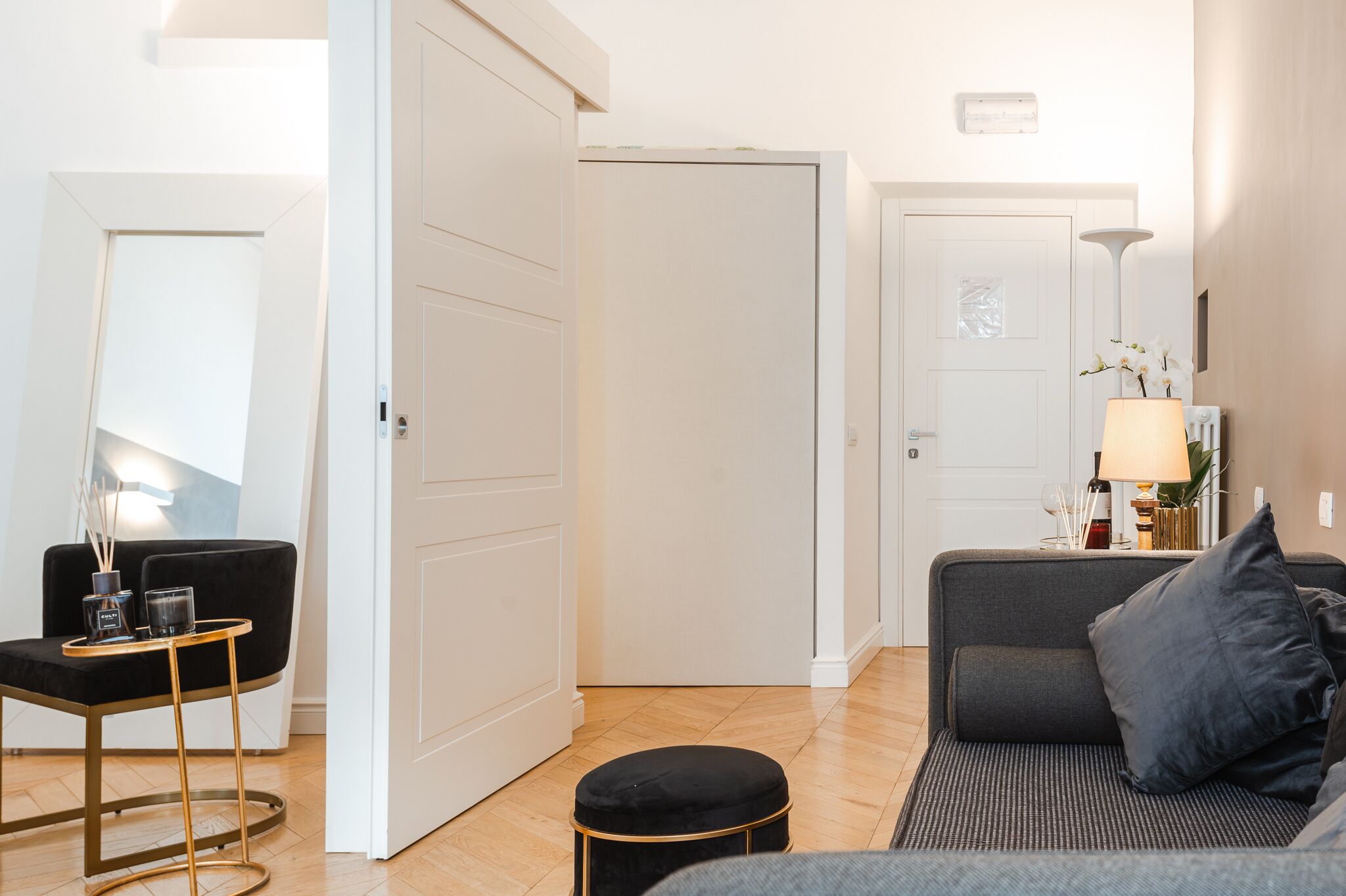 A modern living room with a sofa, a small table with a lamp, and a door leading to another room.