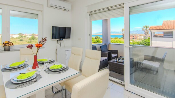 A dining area with a table set for four, a television mounted on the wall, and a view of the sea through a sliding glass door. - Apartment Andreja (Vir)