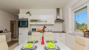 A modern kitchen with white cabinets, a built-in oven, and a microwave. There is a dining table set with plates, glasses, and a red vase with flowers. - Apartment Andreja (Vir)