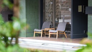 Zona de estar con dos sillas y una mesa frente a un edificio con pared de vidrio que muestra texto repetido.
