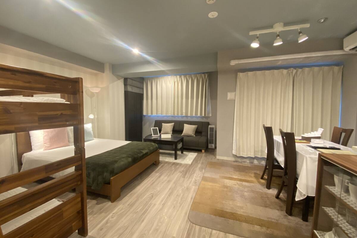A modern hotel room with a bunk bed, a sofa, a dining table with chairs, and a large window with curtains.