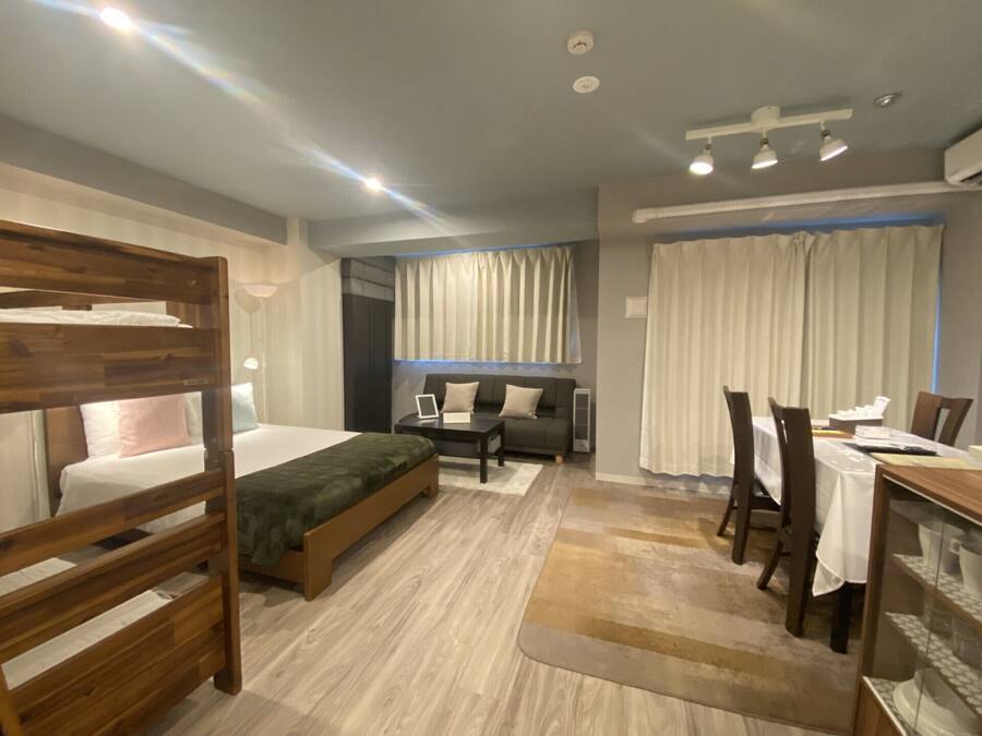 A modern hotel room with a bunk bed, a sofa, a dining table with chairs, and a large window with curtains.