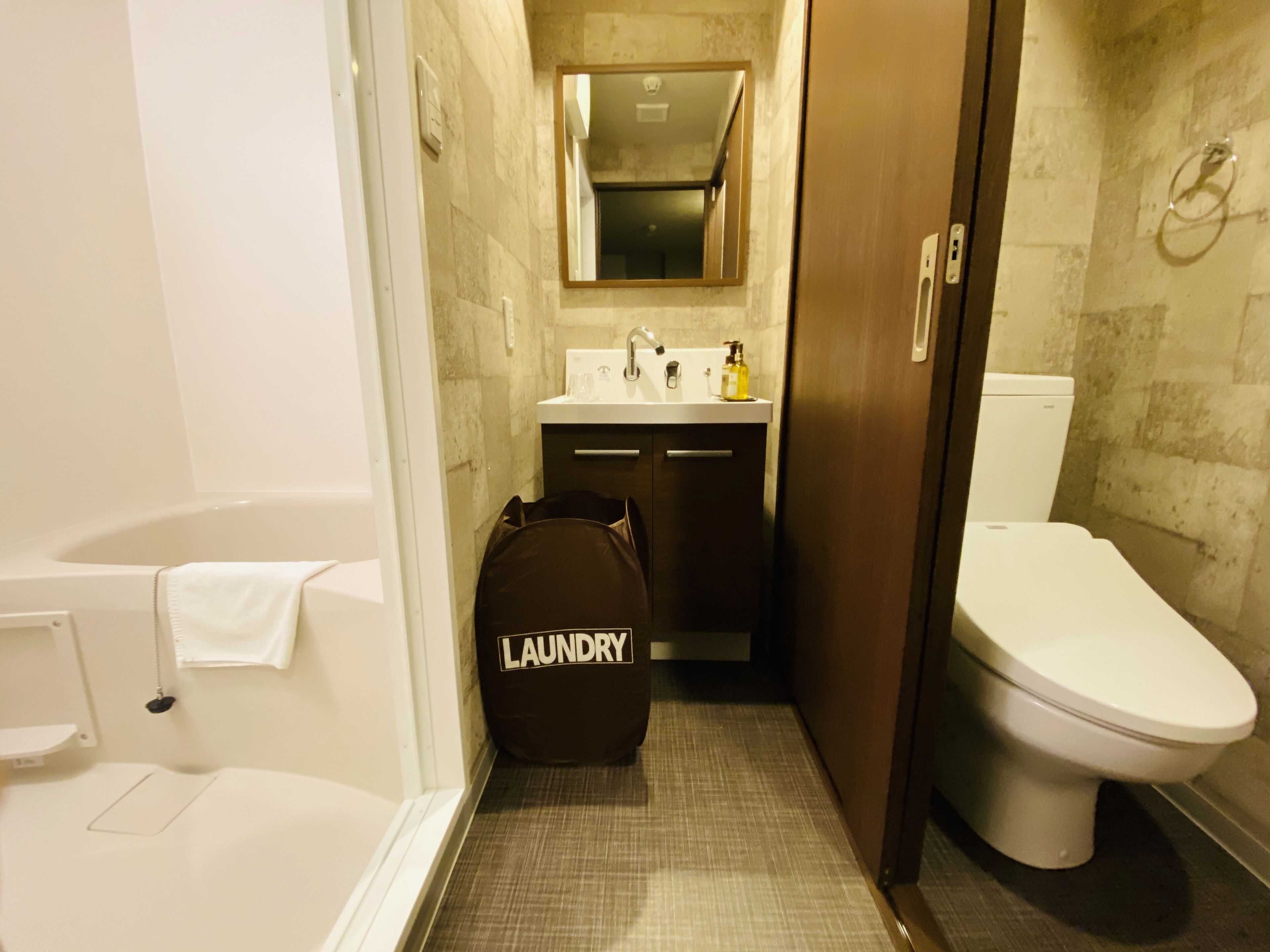 Une petite salle de bain comprenant une baignoire, des toilettes et un lavabo. Un sac à linge est posé sur le sol.