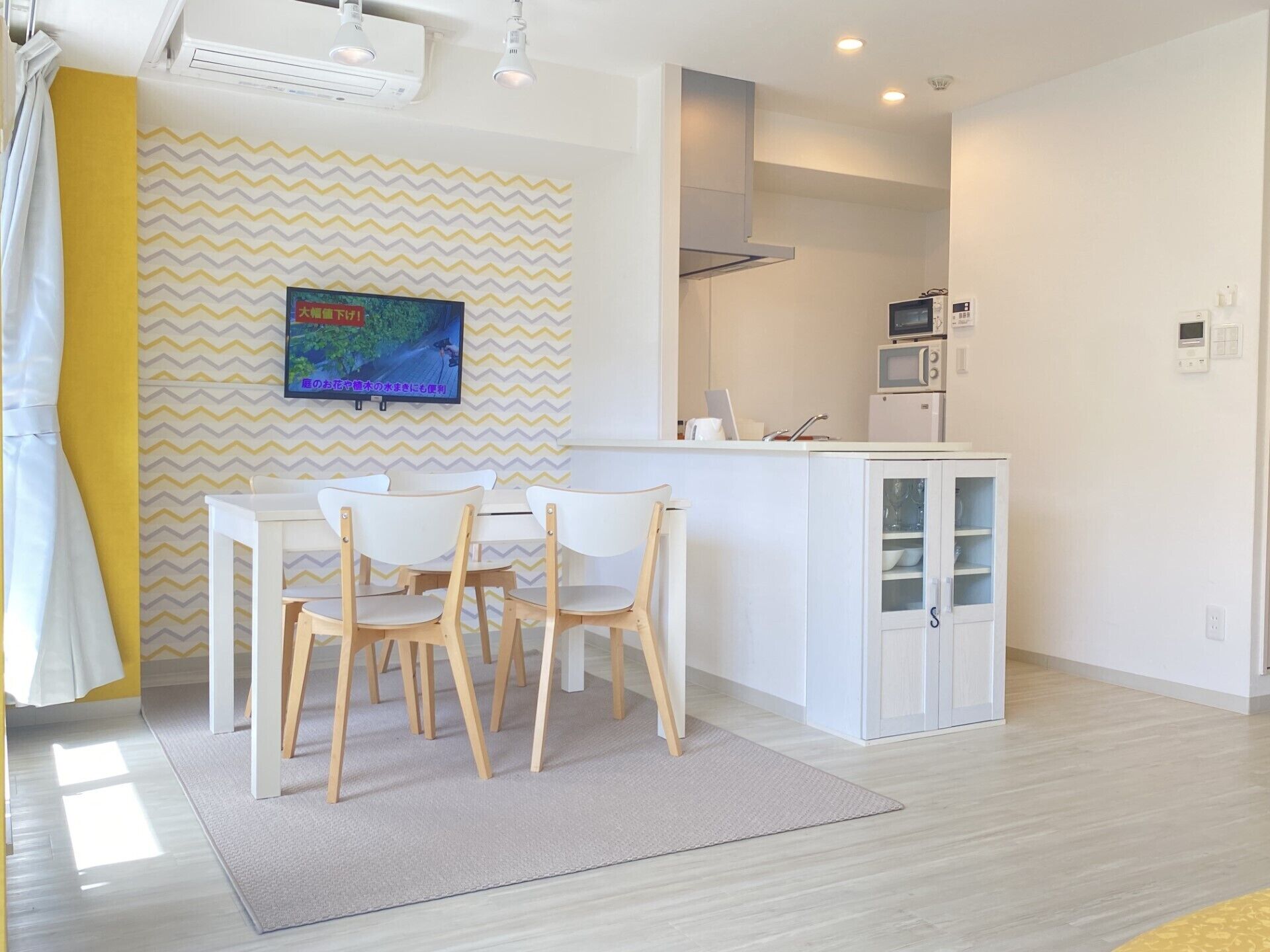 Eine moderne Küche mit weißen Schränken, einer Mikrowelle und einem kleinen Kühlschrank. Es gibt einen Essbereich mit einem weißen Tisch und Stühlen, und ein großes Fernsehgerät an der Wand.