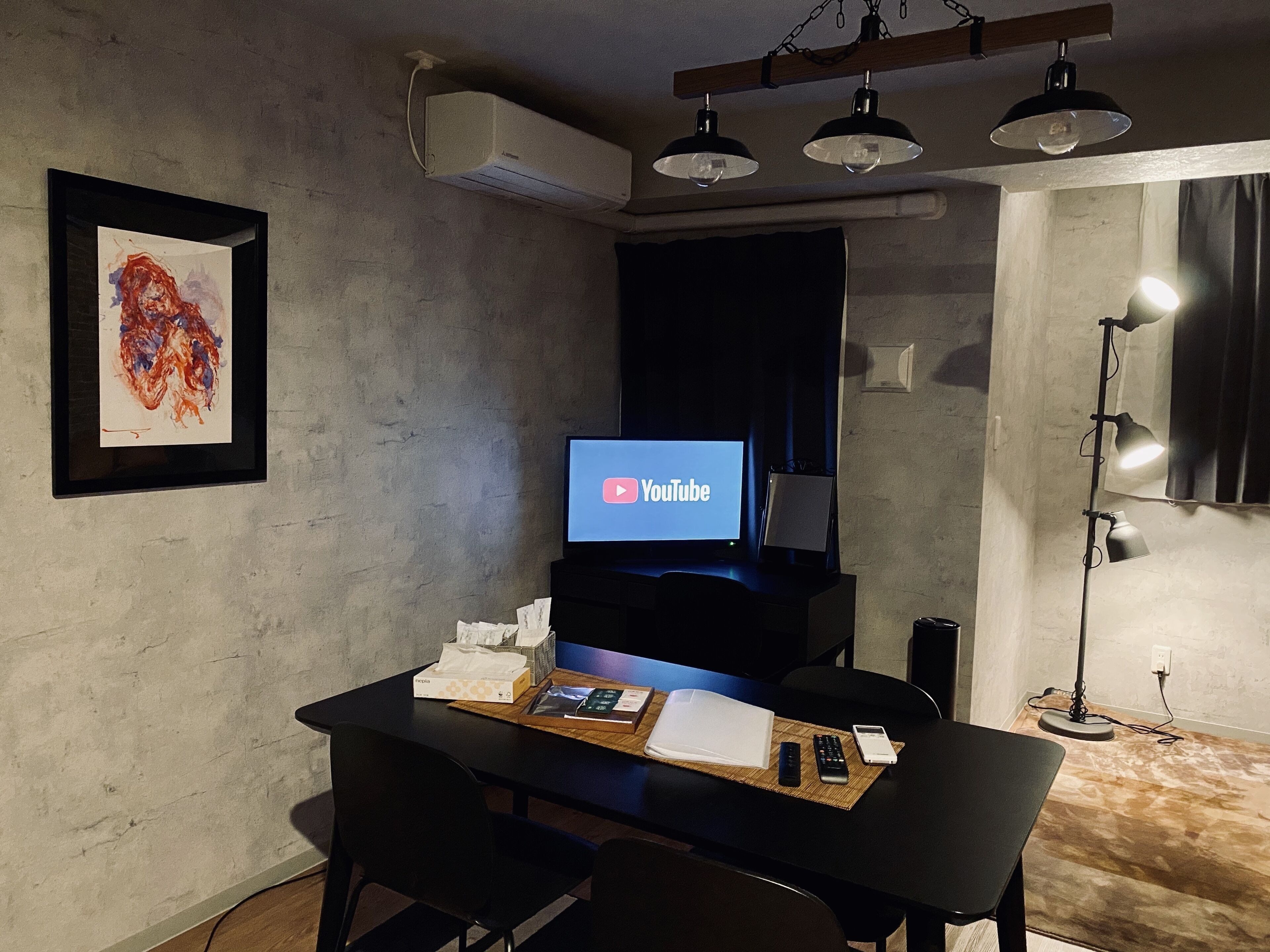 Une pièce comprenant un bureau, une chaise, un téléviseur à écran plat diffusant des vidéos de YouTube, une œuvre d’art encadrée accrochée au mur et une lampe sur pied.