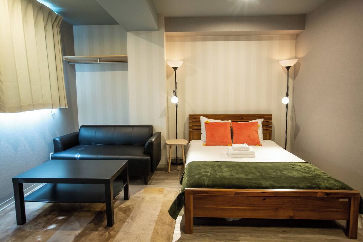A bedroom with a bed, a wooden bench, a black leather sofa, a small table, and two bedside lamps.
