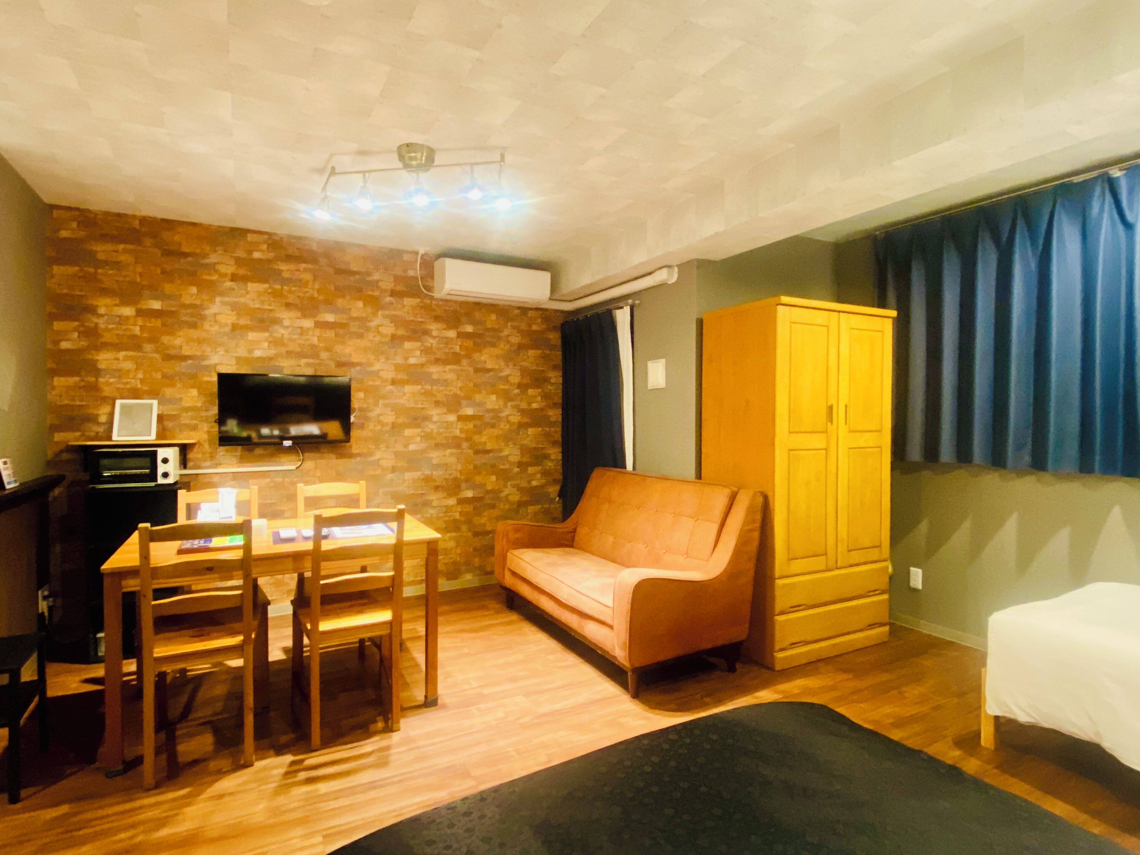 Ein kompakter Wohnraum mit Backsteinwand, einem Flachbildfernseher, einem Holzesstisch mit Stühlen, einer Couch, einem Kleiderschrank und einem Bett.