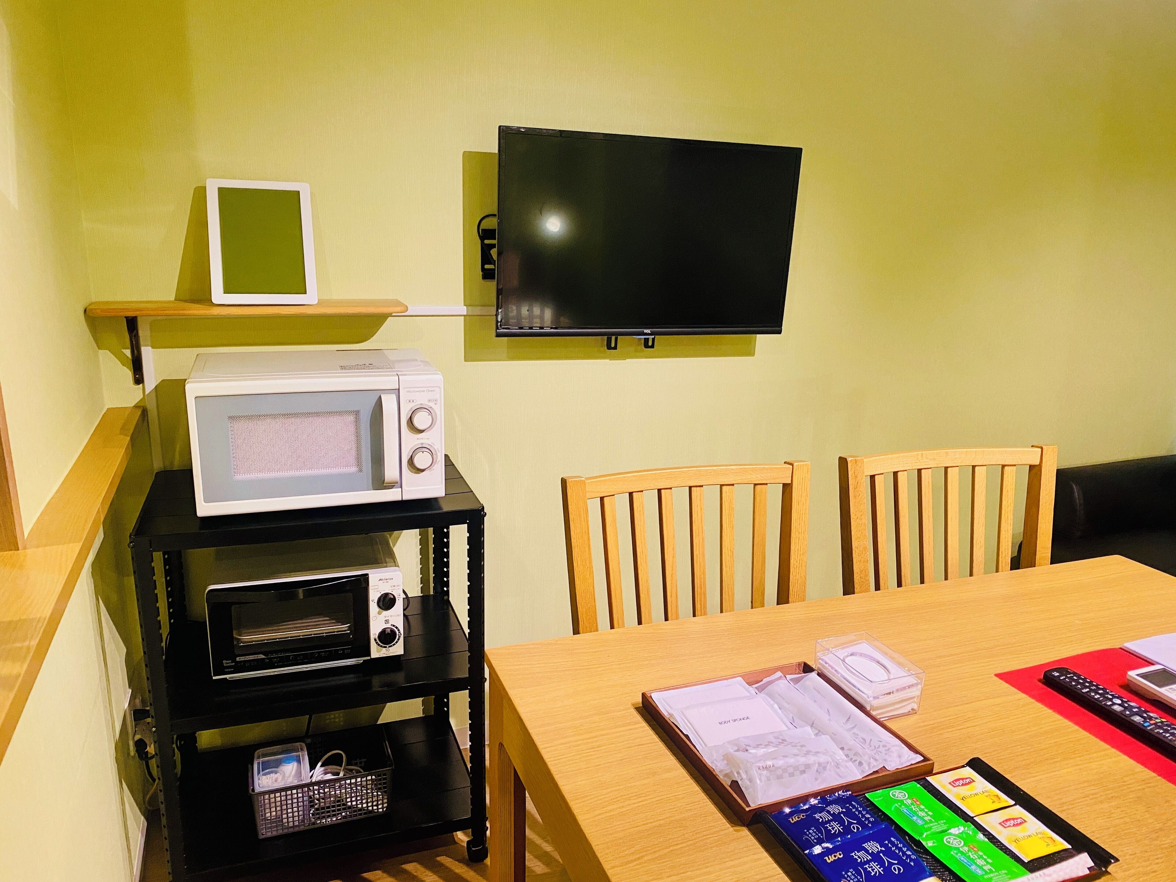 A room with a wooden table, two chairs, a TV mounted on the wall, a microwave on a shelf, and a black TV stand with various items on it.