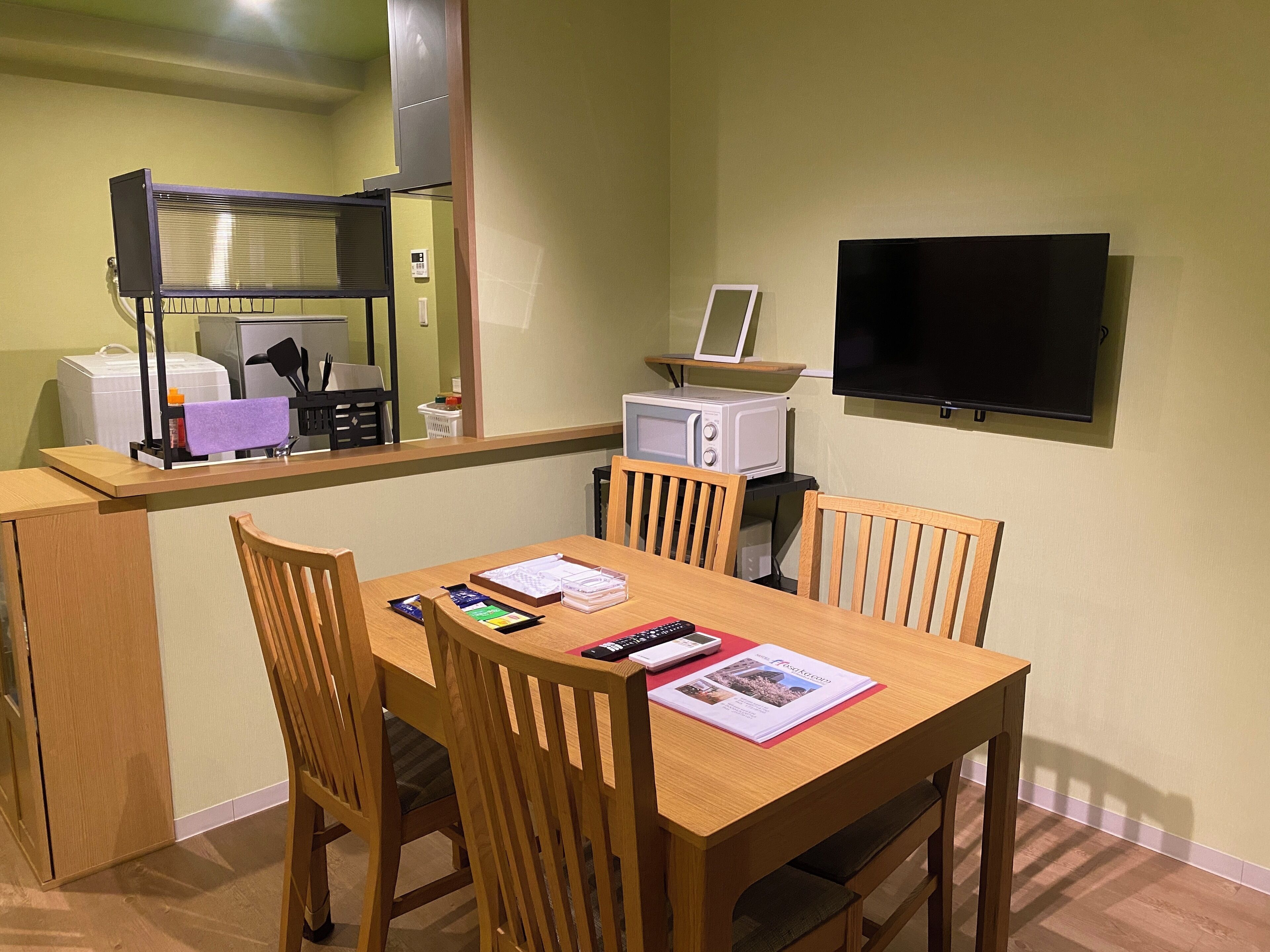 A compact hotel room with a wooden dining table, four chairs, a microwave, a small TV, and a kitchenette area with a refrigerator and a microwave.