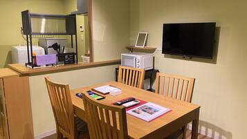 A compact hotel room with a wooden dining table, four chairs, a microwave, a small TV, and a kitchenette area with a refrigerator and a microwave.