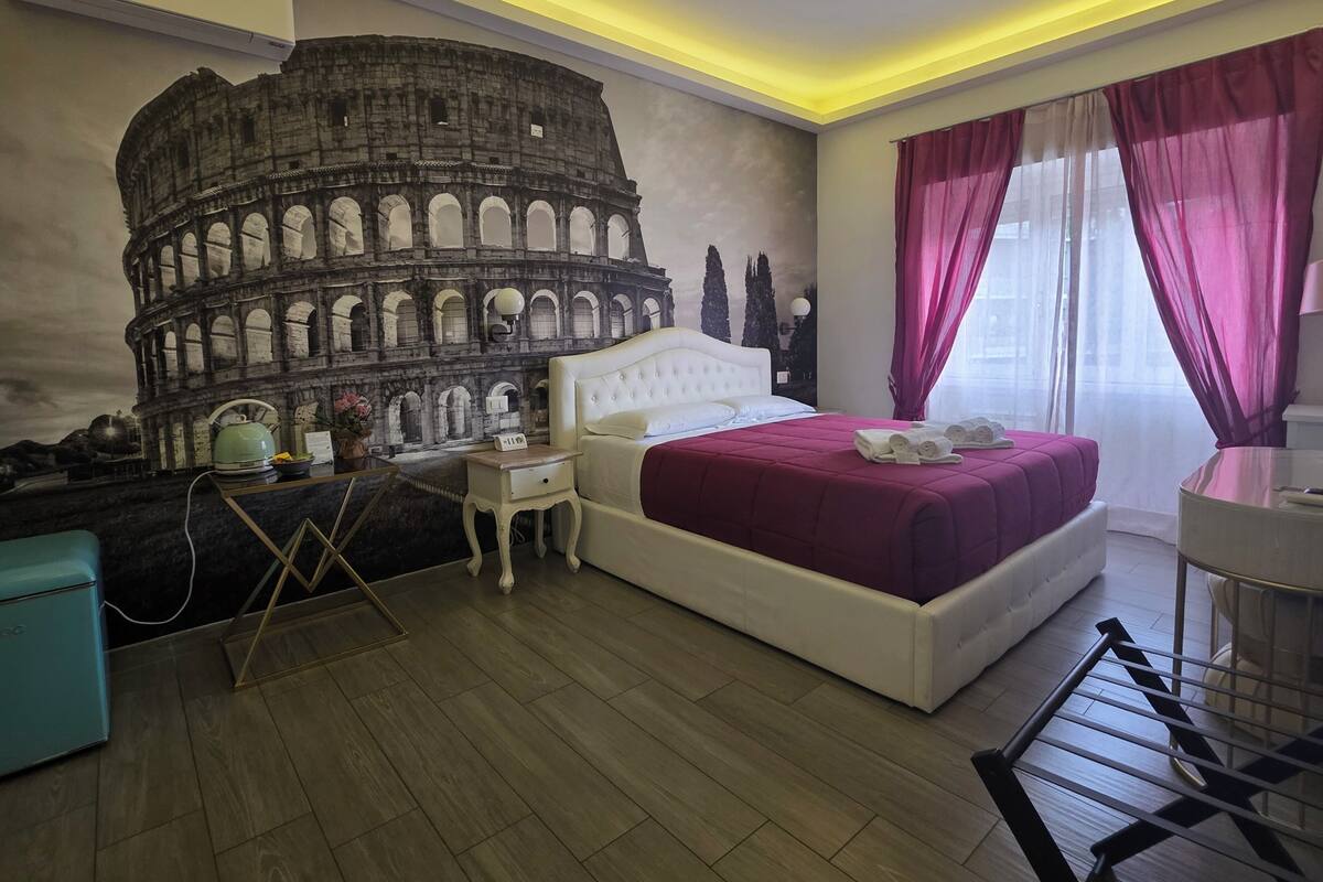 A bedroom with a large mural of the Colosseum, a white bed, a nightstand, and a window with sheer curtains.